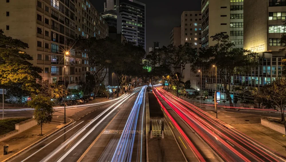 Avenida movimentada à noite com rastros de luz dos veículos entre prédios